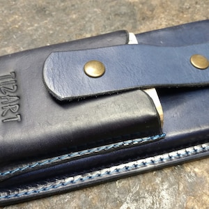May include: A dark blue leather phone case with a flap closure and a brass button. The case has a blue stitching detail and the text "TIZART" embossed on the front.