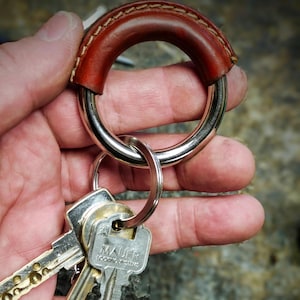 Może przedstawiać: Brązowy skórzany brelok do kluczy z srebrnym metalowym pierścieniem. Pierścień jest przymocowany do kompletu kluczy ze srebrnym łańcuszkiem do kluczy. Klucze mają srebrne wykończenie i są oznaczone "MAUER LOCKING SYSTEMS".