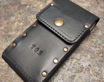 May include: Black leather phone case with brass rivets and a snap closure. The case has a pocket on the front and the initials "TGE" are embossed on the front.