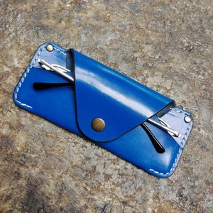 May include: A vibrant blue leather glasses case with white stitching and a brass snap closure. The case holds a pair of black-framed glasses. The case is rectangular and has a flap that folds over to secure the glasses.