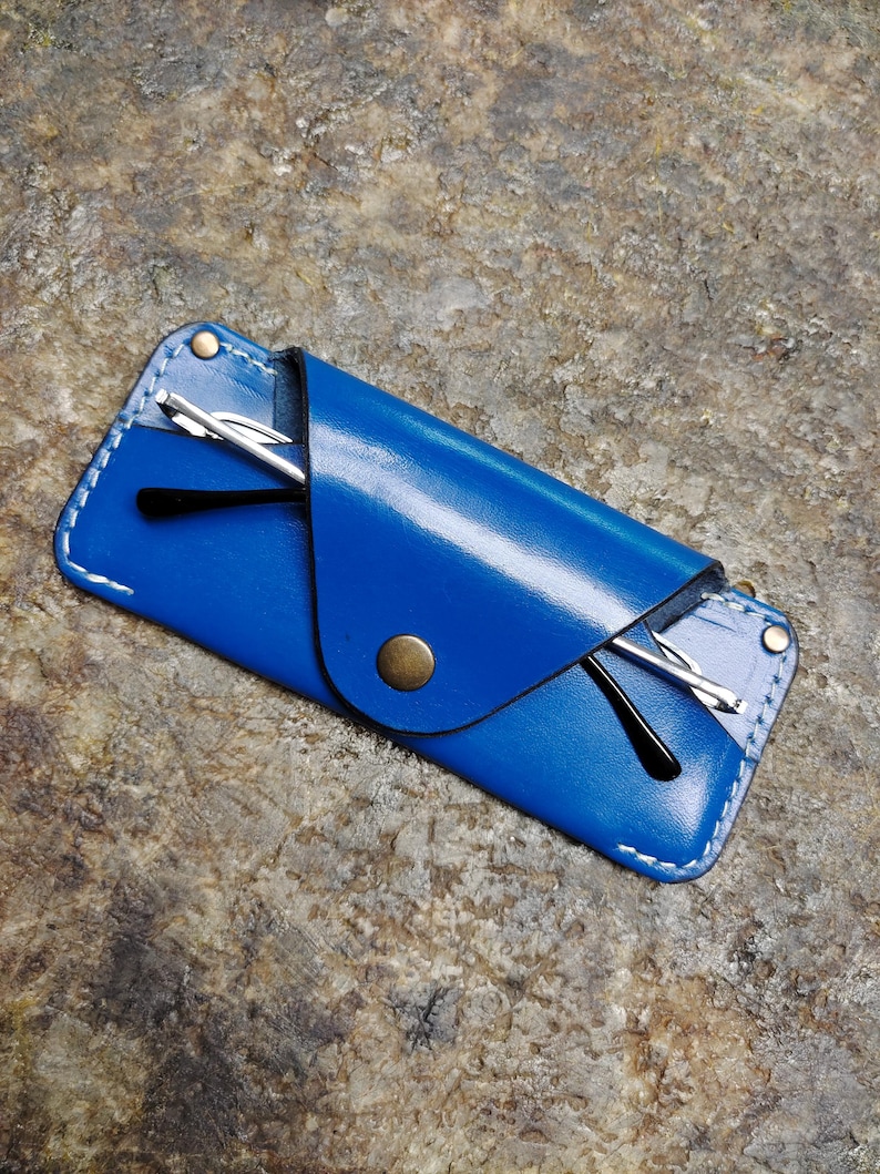 May include: A vibrant blue leather glasses case with white stitching and a brass snap closure. The case holds a pair of black-framed glasses. The case is rectangular and has a flap that folds over to secure the glasses.