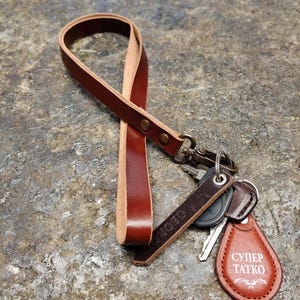 Puede incluir: Una correa de muñeca de cuero marrón con un cierre giratorio de latón y una etiqueta de llavero de cuero en forma de lágrima con el texto blanco "СУПЕР ТАТКО" y adornos, unida a un juego de llaves de metal en una argolla plateada.