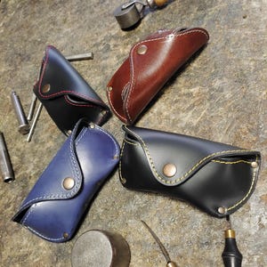 Puede incluir: Cuatro estuches de cuero para gafas en negro, marr&oacute;n, azul y negro, con costuras en contraste y cierres de bot&oacute;n a presi&oacute;n. Los estuches est&aacute;n colocados sobre una superficie texturizada con herramientas de cuero.