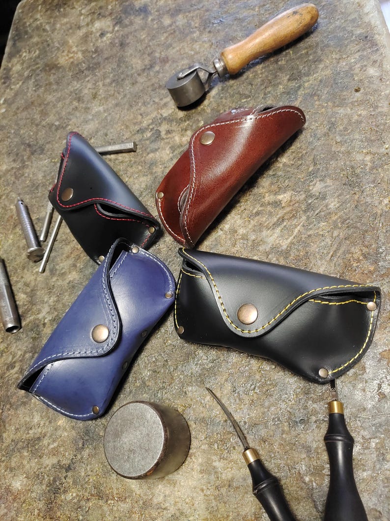 Puede incluir: Cuatro estuches de cuero para gafas en negro, marr&oacute;n, azul y negro, con costuras en contraste y cierres de bot&oacute;n a presi&oacute;n. Los estuches est&aacute;n colocados sobre una superficie texturizada con herramientas de cuero.