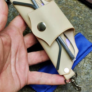 Puede incluir: Estuche de gafas de cuero beige con cierre de bot&oacute;n a presi&oacute;n, que contiene gafas con montura negra. Tambi&eacute;n se ve un pa&ntilde;o de microfibra azul y un mosquet&oacute;n plateado. El estuche est&aacute; dise&ntilde;ado para proteger las gafas.