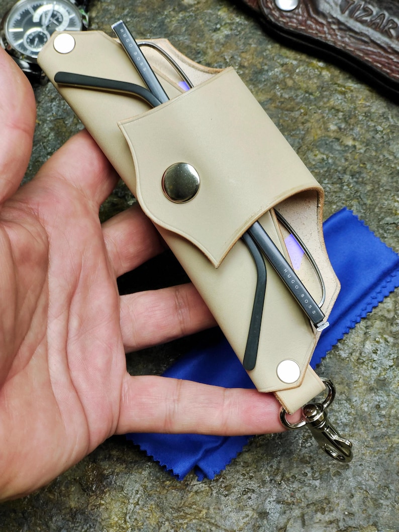 Puede incluir: Estuche de gafas de cuero beige con cierre de bot&oacute;n a presi&oacute;n, que contiene gafas con montura negra. Tambi&eacute;n se ve un pa&ntilde;o de microfibra azul y un mosquet&oacute;n plateado. El estuche est&aacute; dise&ntilde;ado para proteger las gafas.