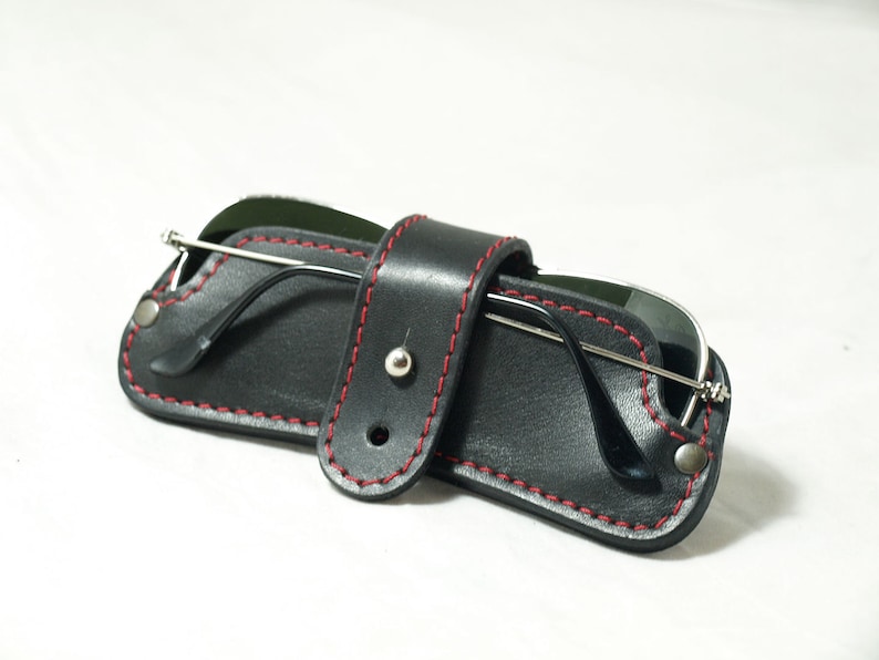 May include: A black leather sunglasses case with red stitching and a silver snap closure. The case is folded closed and the sunglasses are inside.
