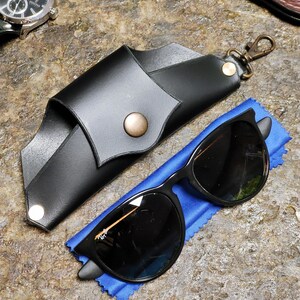 Puede incluir: Gafas de sol negras con un estuche de cuero negro y un pa&ntilde;o de microfibra azul. El estuche tiene un cierre a presi&oacute;n y un cierre met&aacute;lico. Tambi&eacute;n se ve un reloj plateado con una correa de cuero negra.