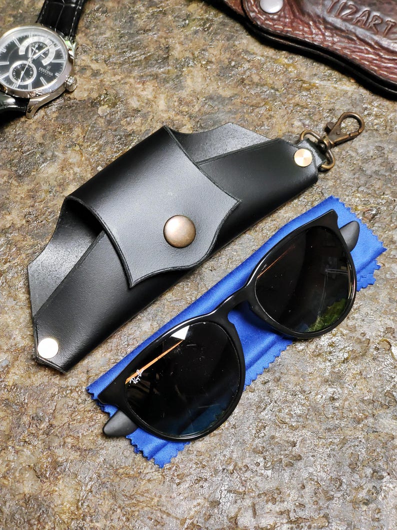 Puede incluir: Gafas de sol negras con un estuche de cuero negro y un pa&ntilde;o de microfibra azul. El estuche tiene un cierre a presi&oacute;n y un cierre met&aacute;lico. Tambi&eacute;n se ve un reloj plateado con una correa de cuero negra.