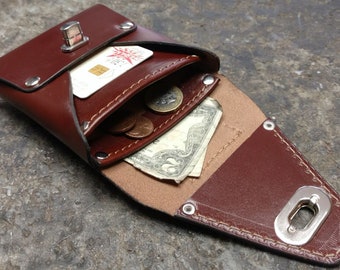 Puede incluir: Una cartera de cuero marrón con cierre de solapa y un broche de metal. La cartera está abierta y muestra una tarjeta de crédito, dinero en efectivo y monedas en su interior.