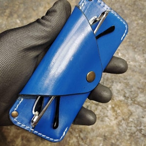 May include: A vibrant blue leather glasses case with white stitching and a brass snap closure. The case holds a pair of eyeglasses with black frames and silver arms. The case is designed to protect eyewear.
