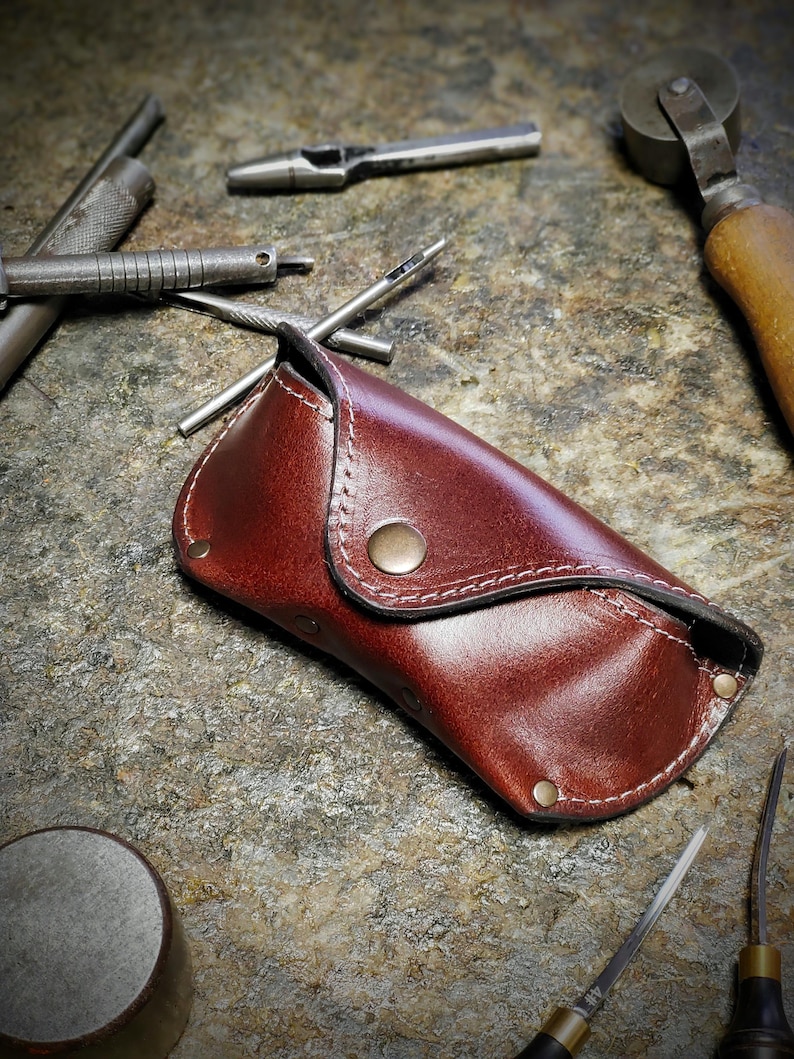 Puede incluir: Un estuche de gafas de cuero marr&oacute;n rojizo, cosido a mano con hilo blanco y un cierre de bot&oacute;n de lat&oacute;n. Varias herramientas met&aacute;licas, como punzones y agujas, sugieren un taller de artesan&iacute;a en cuero.