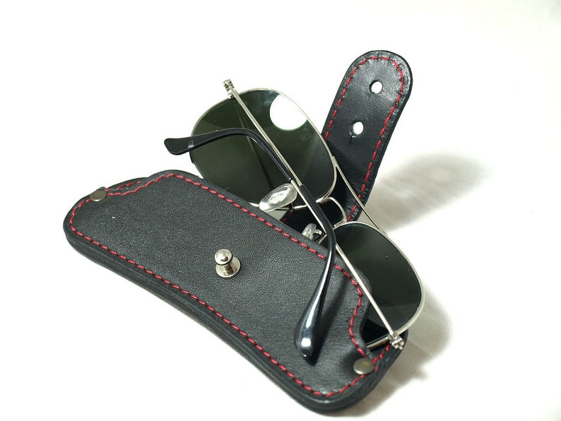 K&ouml;nnte beinhalten: Ein schwarzes Lederetui f&uuml;r Sonnenbrillen mit roten N&auml;hten und einem silbernen Druckknopfverschluss. Das Etui ist ge&ouml;ffnet und eine Sonnenbrille befindet sich darin.