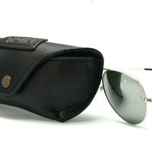 K&ouml;nnte beinhalten: Eine silberne Aviator-Sonnenbrille mit dunkelgrauen Gl&auml;sern und einem schwarzen Lederetui. Das Etui hat ein braunes Leder-Etikett mit dem gepr&auml;gten Text "The Factory".