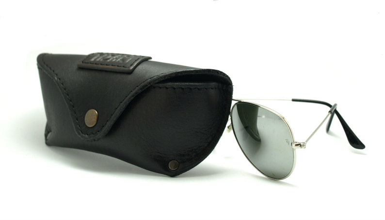 K&ouml;nnte beinhalten: Eine silberne Aviator-Sonnenbrille mit dunkelgrauen Gl&auml;sern und einem schwarzen Lederetui. Das Etui hat ein braunes Leder-Etikett mit dem gepr&auml;gten Text "The Factory".