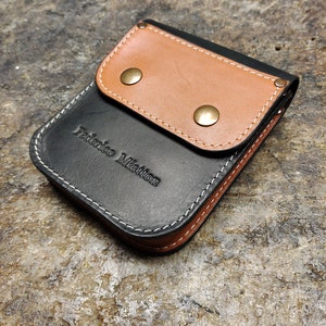 Puede incluir: Una cartera de cuero negro con una solapa de cuero marrón y dos broches de latón. La cartera tiene un detalle de costura blanca y el nombre "Federico Miatton" grabado en la parte delantera.