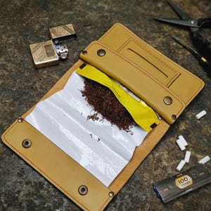May include: Tan leather tobacco pouch with red stitching, open to reveal a packet of tobacco. A gold-colored lighter, scissors, and a pack of rolling papers are also visible. Several white filters are scattered around the pouch.