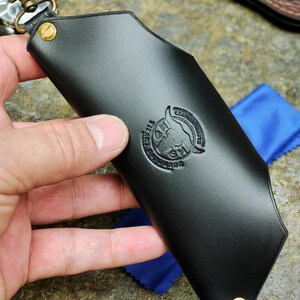 Puede incluir: Un llavero de cuero negro con un mosquet&oacute;n y remaches de lat&oacute;n. El llavero tiene un logotipo en relieve y est&aacute; sujeto por una mano. Un pa&ntilde;o azul es visible en el fondo.