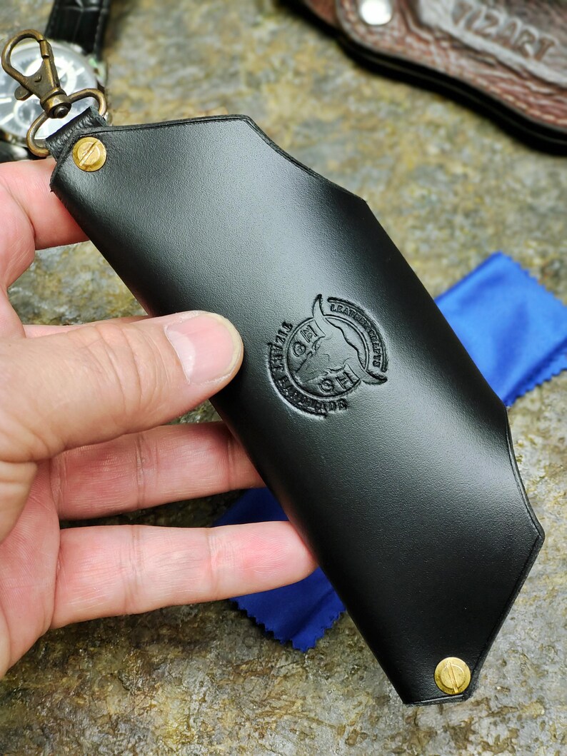 Puede incluir: Un llavero de cuero negro con un mosquet&oacute;n y remaches de lat&oacute;n. El llavero tiene un logotipo en relieve y est&aacute; sujeto por una mano. Un pa&ntilde;o azul es visible en el fondo.