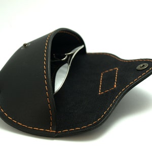 Puede incluir: Estuche de gafas de cuero negro con costuras naranjas y cierre de bot&oacute;n a presi&oacute;n. El estuche est&aacute; abierto, revelando un par de gafas de sol. El estuche tiene una costura decorativa en forma de diamante.