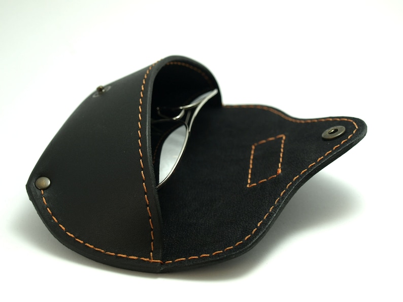 Puede incluir: Estuche de gafas de cuero negro con costuras naranjas y cierre de bot&oacute;n a presi&oacute;n. El estuche est&aacute; abierto, revelando un par de gafas de sol. El estuche tiene una costura decorativa en forma de diamante.