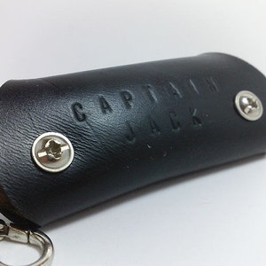 May include: Black leather key chain with the words "Captain Jack" embossed on the front. The keychain has two silver screws.