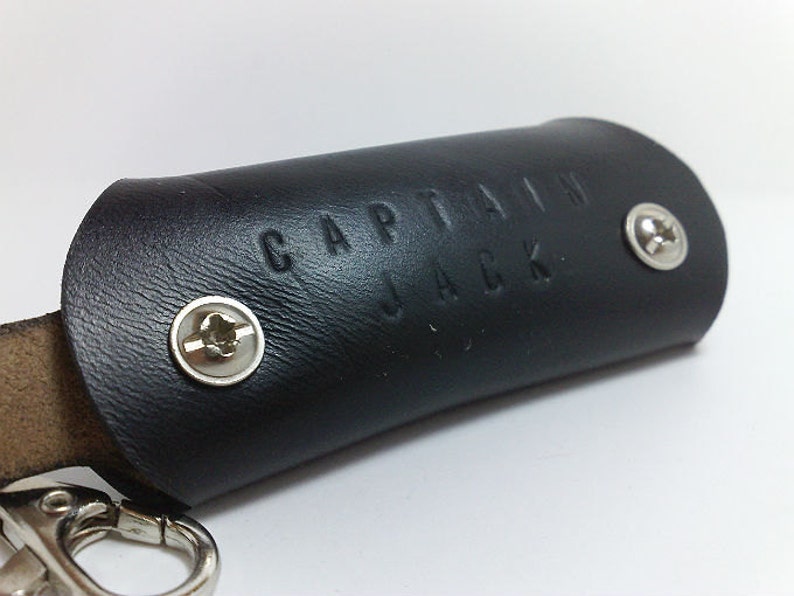 May include: Black leather key chain with the words "Captain Jack" embossed on the front. The keychain has two silver screws.