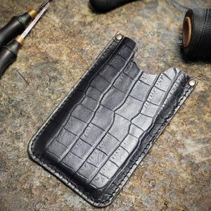 May include: A black leather phone case with a crocodile pattern. The case has a stitched edge and is lying on a textured surface.