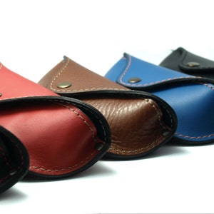 Puede incluir: Una fila de estuches de cuero para gafas de colores: negro, rojo, marr&oacute;n, azul y negro. Cada estuche tiene un cierre de bot&oacute;n a presi&oacute;n y costuras en contraste. Los estuches est&aacute;n dispuestos en diagonal sobre un fondo blanco.