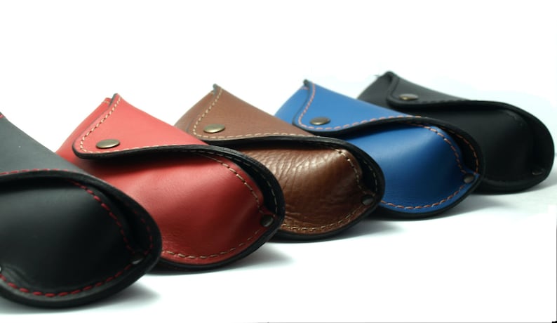 Puede incluir: Una fila de estuches de cuero para gafas de colores: negro, rojo, marr&oacute;n, azul y negro. Cada estuche tiene un cierre de bot&oacute;n a presi&oacute;n y costuras en contraste. Los estuches est&aacute;n dispuestos en diagonal sobre un fondo blanco.