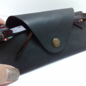 May include: A black leather glasses case with red stitching and a brass button closure. The case holds a pair of eyeglasses with brown frames. The case is designed to protect and store eyewear.