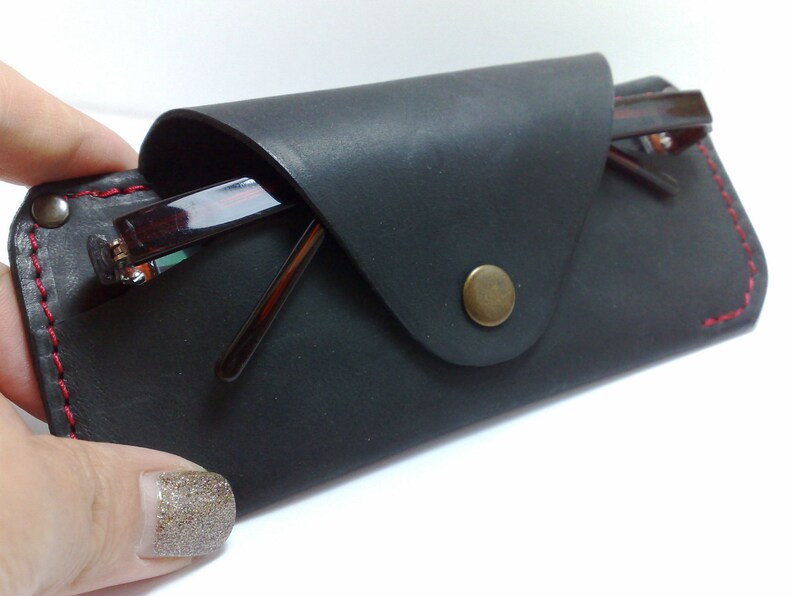 May include: A black leather glasses case with red stitching and a brass button closure. The case holds a pair of eyeglasses with brown frames. The case is designed to protect and store eyewear.