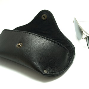 K&ouml;nnte beinhalten: Ein schwarzes Lederetui f&uuml;r Sonnenbrillen mit einem Druckknopfverschluss. Das Etui ist ge&ouml;ffnet und eine silberne Aviator-Sonnenbrille mit verspiegelten Gl&auml;sern liegt daneben.