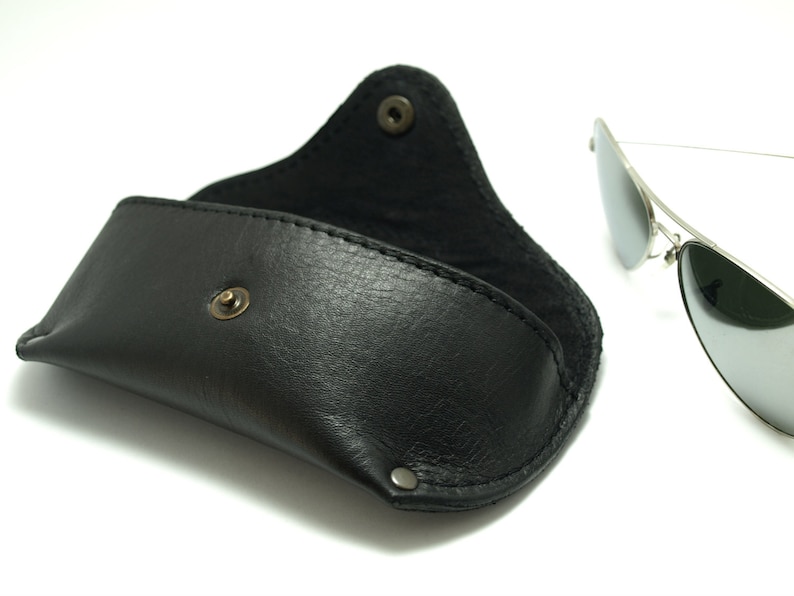 K&ouml;nnte beinhalten: Ein schwarzes Lederetui f&uuml;r Sonnenbrillen mit einem Druckknopfverschluss. Das Etui ist ge&ouml;ffnet und eine silberne Aviator-Sonnenbrille mit verspiegelten Gl&auml;sern liegt daneben.