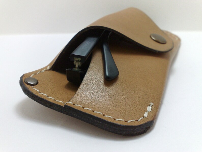May include: A tan leather glasses case with a snap closure, holding a pair of black-framed glasses. The case is stitched with white thread and has a dark brown edge.