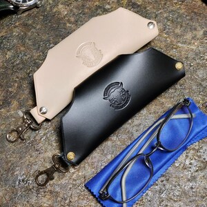 Puede incluir: Dos estuches de cuero para gafas, uno negro y otro beige, con cierres met&aacute;licos y un logotipo. Un par de gafas descansa sobre un pa&ntilde;o azul junto al estuche negro. Se ve un reloj de pulsera.