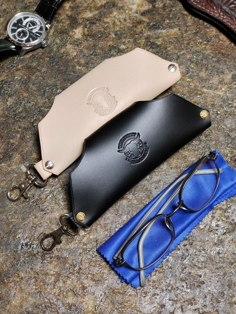 Puede incluir: Dos estuches de cuero para gafas, uno negro y otro beige, con cierres met&aacute;licos y un logotipo. Un par de gafas descansa sobre un pa&ntilde;o azul junto al estuche negro. Se ve un reloj de pulsera.