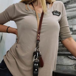 Könnte beinhalten: Ein langes, zweifarbiges Leder-Lanyard mit einem dunkelbraunen Riemen und einer hellbraunen Unterseite. Das Lanyard verfügt über goldfarbene Metallnieten, einen drehbaren Karabinerhaken und einen silberfarbenen Metallring mit Schlüsseln.