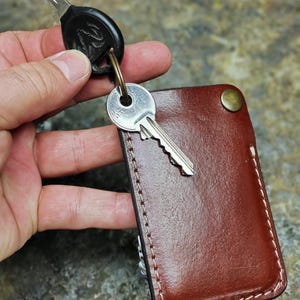 Op de afbeelding: Een bruine leren sleutelhouder met witte stiksels en een messing knoop. Twee sleutels zijn met een metalen ring aan de houder bevestigd. De rechthoekige sleutelhouder lijkt handgemaakt.