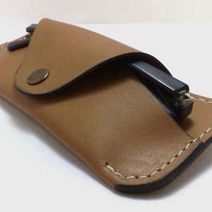 May include: A tan leather glasses case with a flap closure and a metal snap. The case is open, revealing a pair of black glasses. The edges are stitched with white thread, and the case has a minimalist design.
