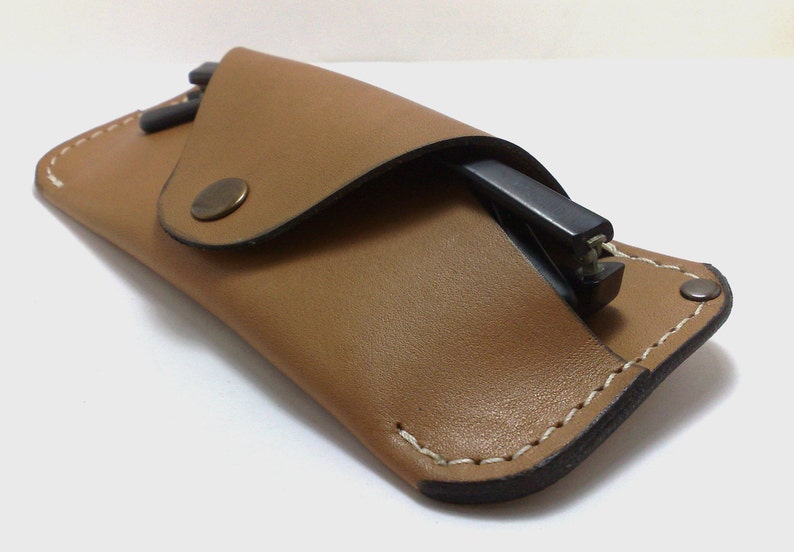 May include: A tan leather glasses case with a flap closure and a metal snap. The case is open, revealing a pair of black glasses. The edges are stitched with white thread, and the case has a minimalist design.