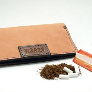 May include: A tan leather wallet with blue stitching and the embossed text "TIZART". The wallet is next to a pile of tobacco, several white filters, and a pack of rolling papers.
