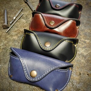Puede incluir: Cuatro estuches de cuero para gafas en negro, marr&oacute;n y azul, cada uno con costuras en contraste y cierre de bot&oacute;n a presi&oacute;n. Los estuches est&aacute;n apilados, mostrando los diferentes colores y detalles de costura. Se ven herramientas en el fondo.
