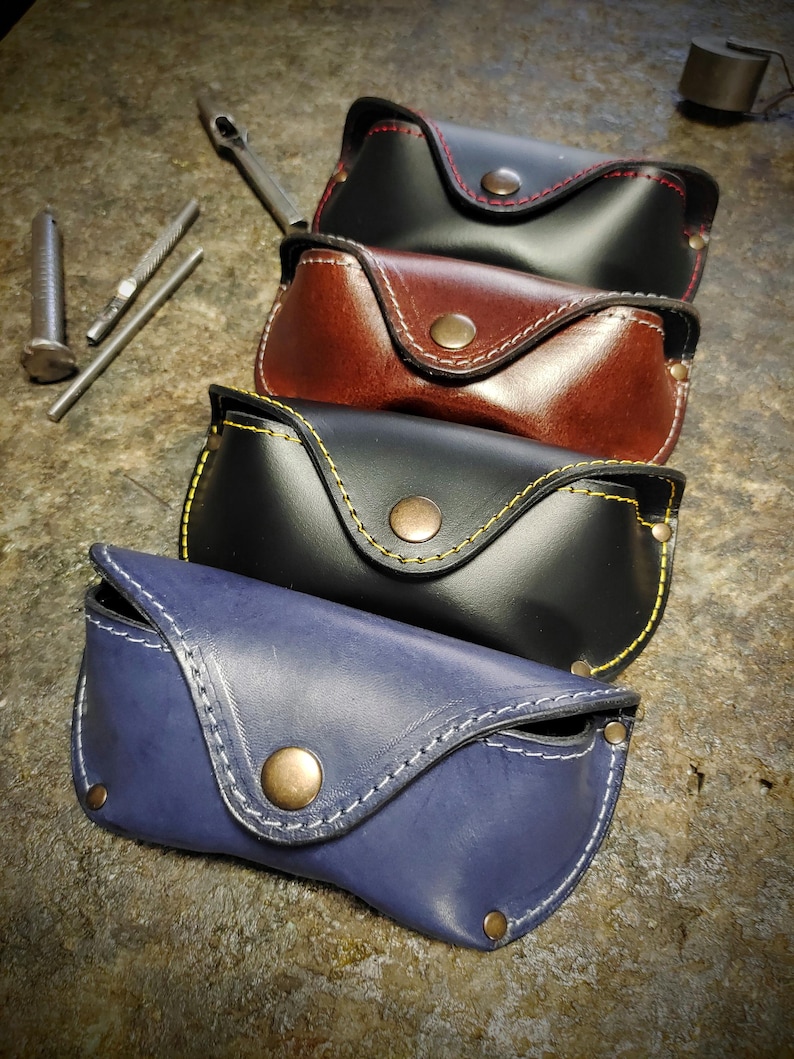 Puede incluir: Cuatro estuches de cuero para gafas en negro, marr&oacute;n y azul, cada uno con costuras en contraste y cierre de bot&oacute;n a presi&oacute;n. Los estuches est&aacute;n apilados, mostrando los diferentes colores y detalles de costura. Se ven herramientas en el fondo.