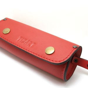 May include: Red leather pencil case with a zipper closure and a metal clasp. The case has a small strap with a snap closure and the word "tizart" embossed on the front.