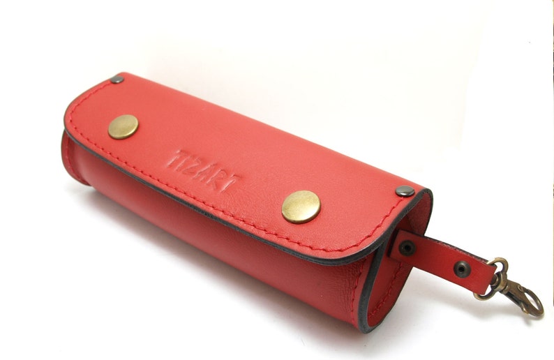 May include: Red leather pencil case with a zipper closure and a metal clasp. The case has a small strap with a snap closure and the word "tizart" embossed on the front.
