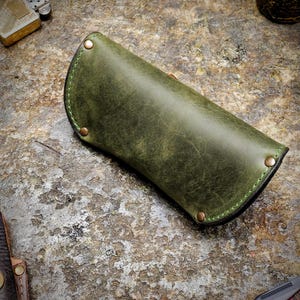 Puede incluir: Un estuche para gafas de cuero verde oliva hecho a mano, con remaches de cobre y costuras verdes. El estuche tiene un dise&ntilde;o curvo y un acabado envejecido. Otros art&iacute;culos en la imagen incluyen un encendedor y una navaja plegable.