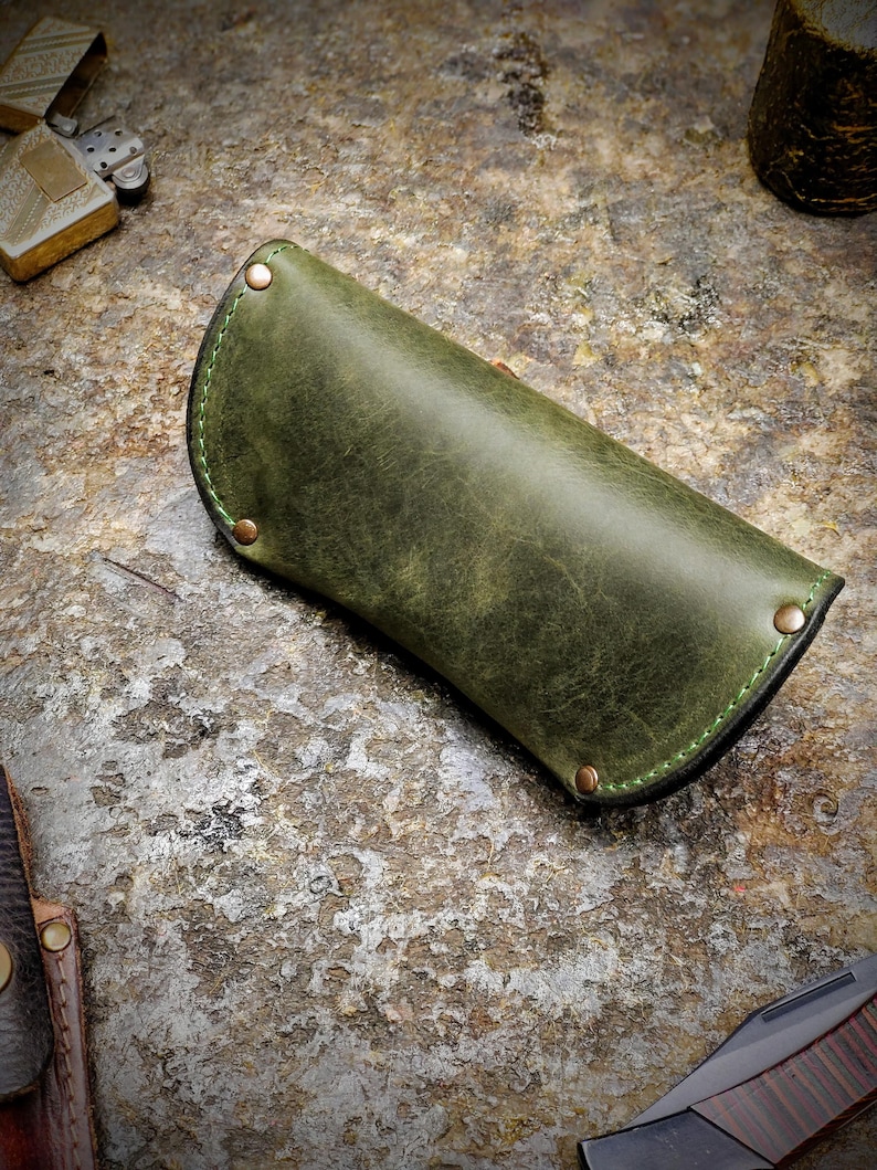 Puede incluir: Un estuche para gafas de cuero verde oliva hecho a mano, con remaches de cobre y costuras verdes. El estuche tiene un dise&ntilde;o curvo y un acabado envejecido. Otros art&iacute;culos en la imagen incluyen un encendedor y una navaja plegable.