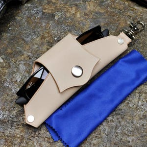 Puede incluir: Funda de gafas de sol de cuero beige con cierre de bot&oacute;n a presi&oacute;n, que contiene gafas de sol negras. Tambi&eacute;n se ve un pa&ntilde;o de limpieza de microfibra azul y un reloj plateado. La funda tiene un cierre plateado.