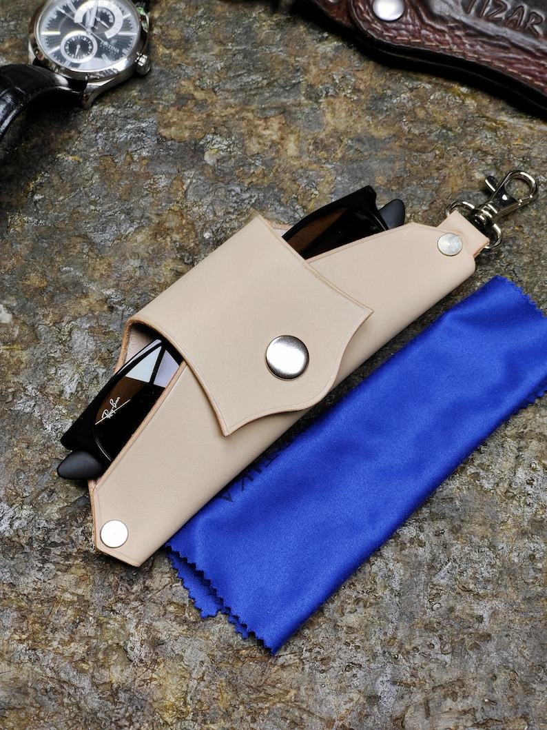Puede incluir: Funda de gafas de sol de cuero beige con cierre de bot&oacute;n a presi&oacute;n, que contiene gafas de sol negras. Tambi&eacute;n se ve un pa&ntilde;o de limpieza de microfibra azul y un reloj plateado. La funda tiene un cierre plateado.